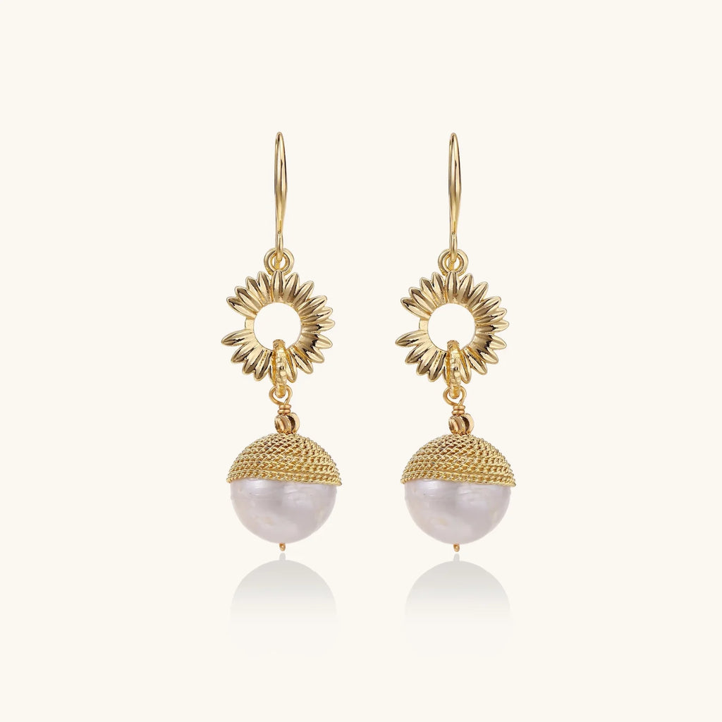 Gold sunburst charm earrings with baroque pearl drops on French hooks against cream background