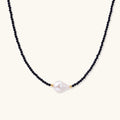 Delicate necklace with black blue sandstone beads and baroque pearl center on white background with gold accents.
