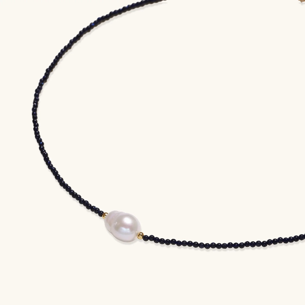 Delicate necklace with black blue sandstone beads and baroque pearl center on white background with gold accents.