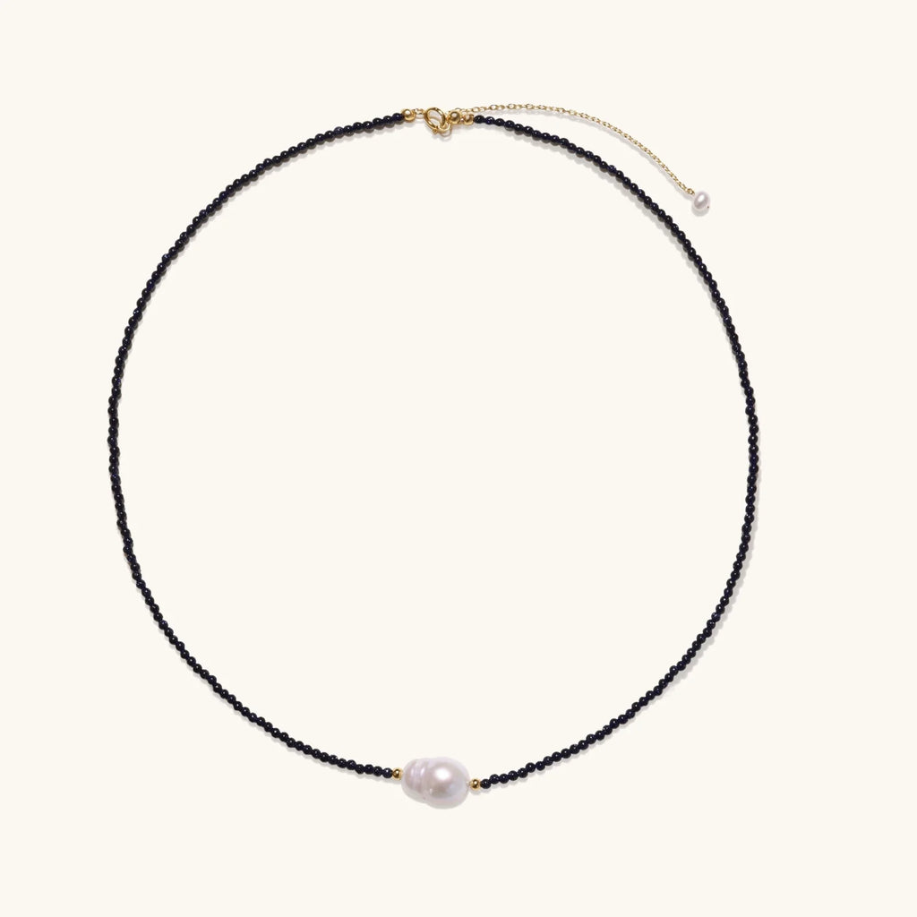 Delicate necklace with black blue sandstone beads and baroque pearl center on white background with gold accents.