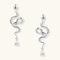 Silver snake-shaped drop earrings with crystal accents and teardrop stone dangles on cream background.