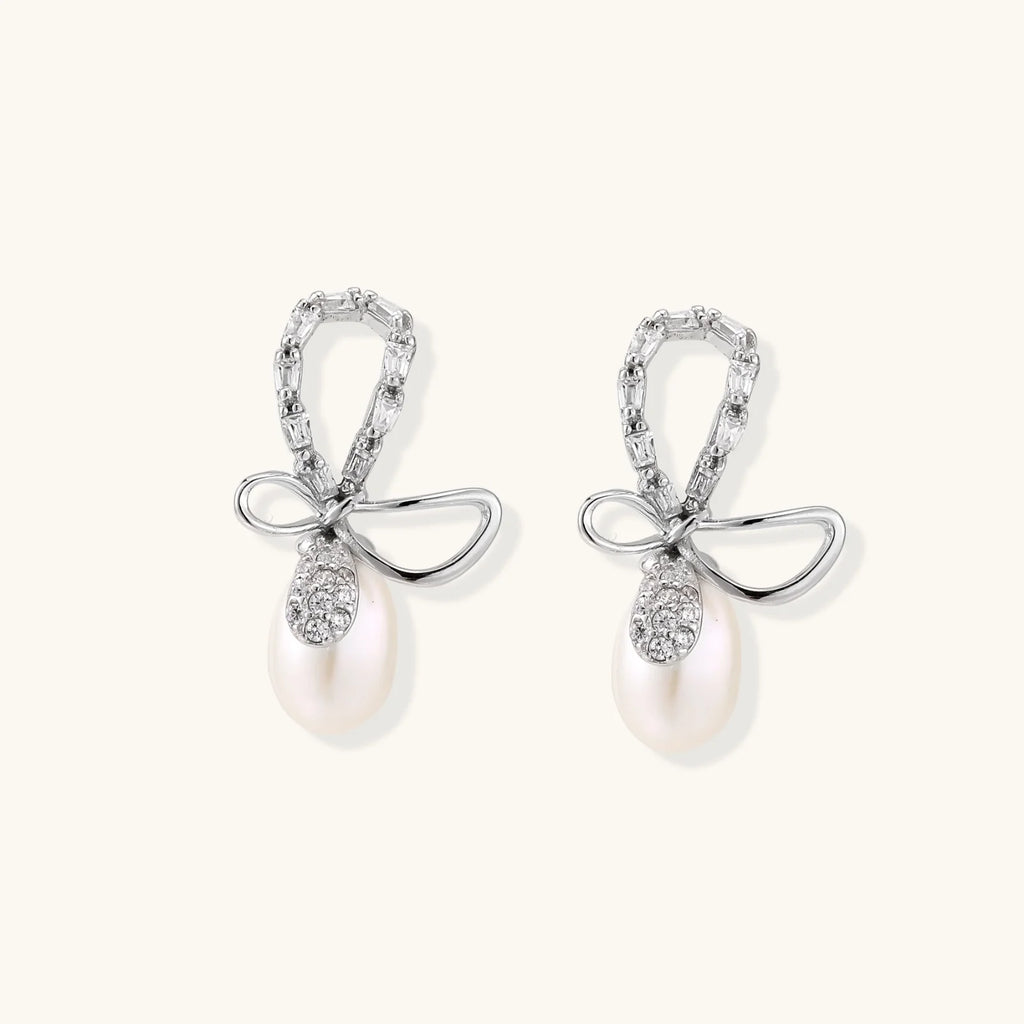 Silver bow-shaped stud earrings with baguette crystals and dangling white freshwater pearls on cream background