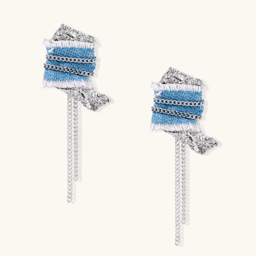Blue denim fabric earrings with white fringe, silver chains, metallic accents, and dangling chain tassels on cream background.