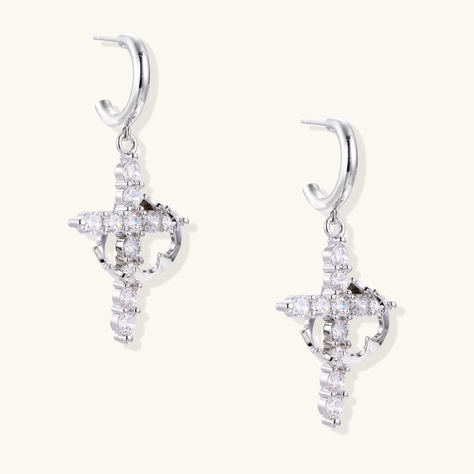 White gold-plated hoop earrings with sparkling zirconia cross charms dangling against cream background