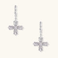 Ornate white gold-plated cross earrings with baguette-cut zirconia stones and twisted leverback hooks on beige background