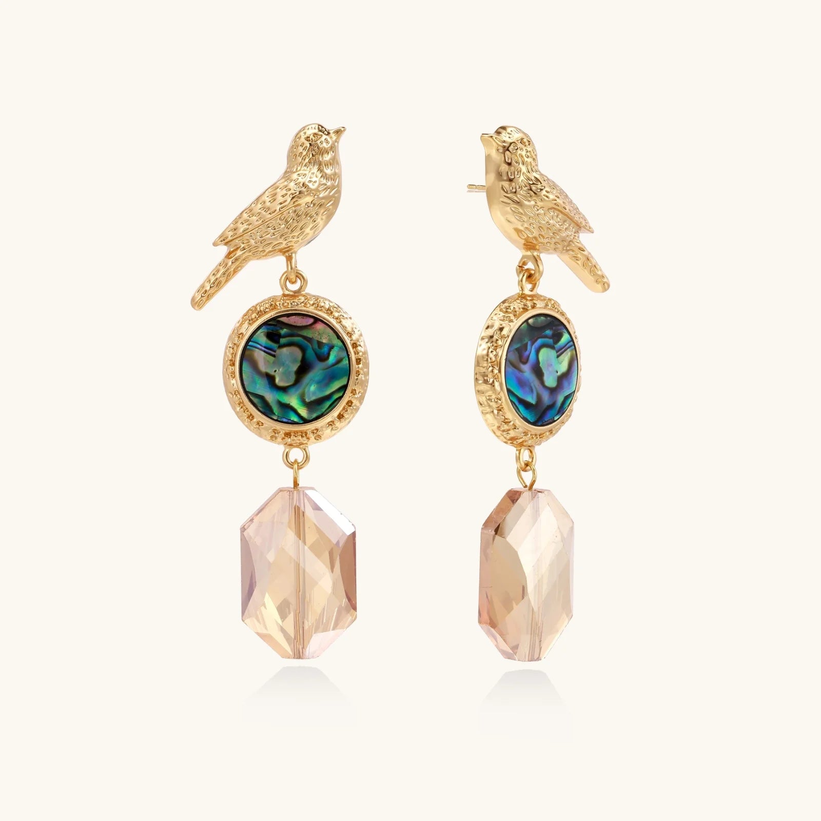 Gold-plated bird stud earrings with iridescent abalone shell medallions and champagne crystal drops on cream background