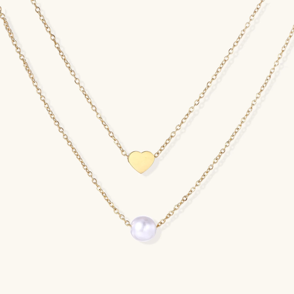 Two delicate gold-plated chains with heart charm and white pearl bead on cream background, minimal layered design.