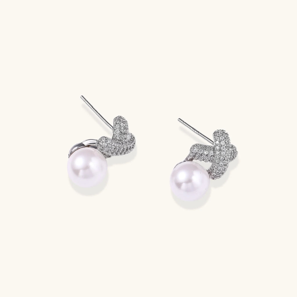 White gold plated studs featuring pavé X-shaped tops with freshwater pearl drops on cream background.