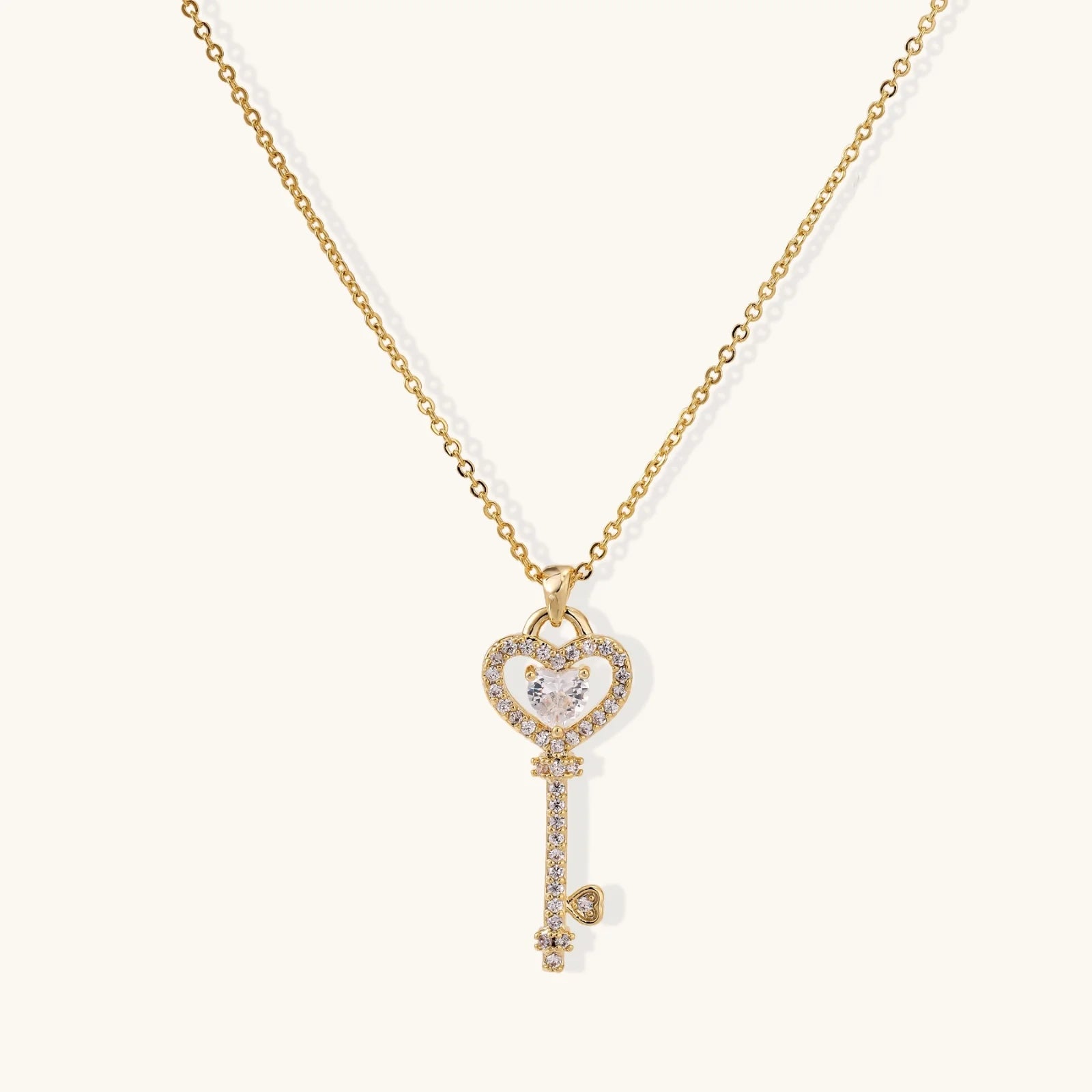 Delicate gold key pendant with heart-shaped bow and crystal pavé detailing on fine chain against cream background.