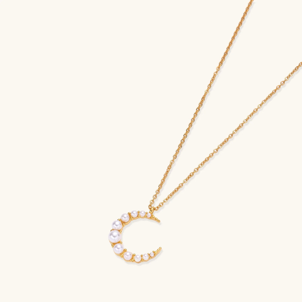 Gold crescent moon pendant with graduated white pearls on delicate chain against cream background