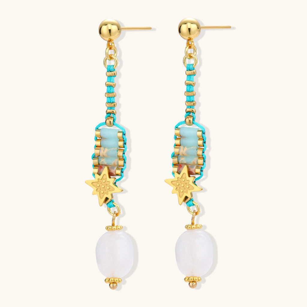 Gold-plated dangle earrings with turquoise threading, star charms, and white pearl beads against cream background.