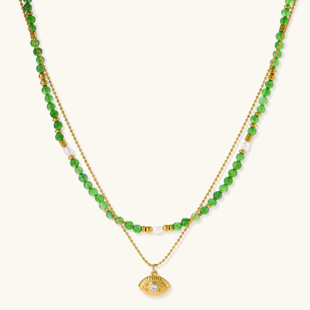Layered necklace with green stone beads, gold spacers, white accents, and protective eye pendant on cream background.